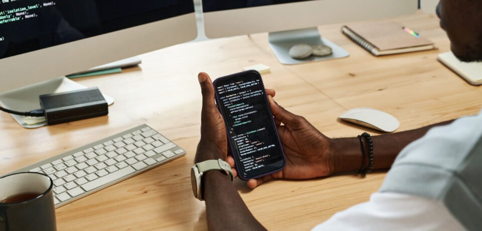 Hands of young black man scrolling through codes in smartphone while sitting by workplace in front of computer screens with data