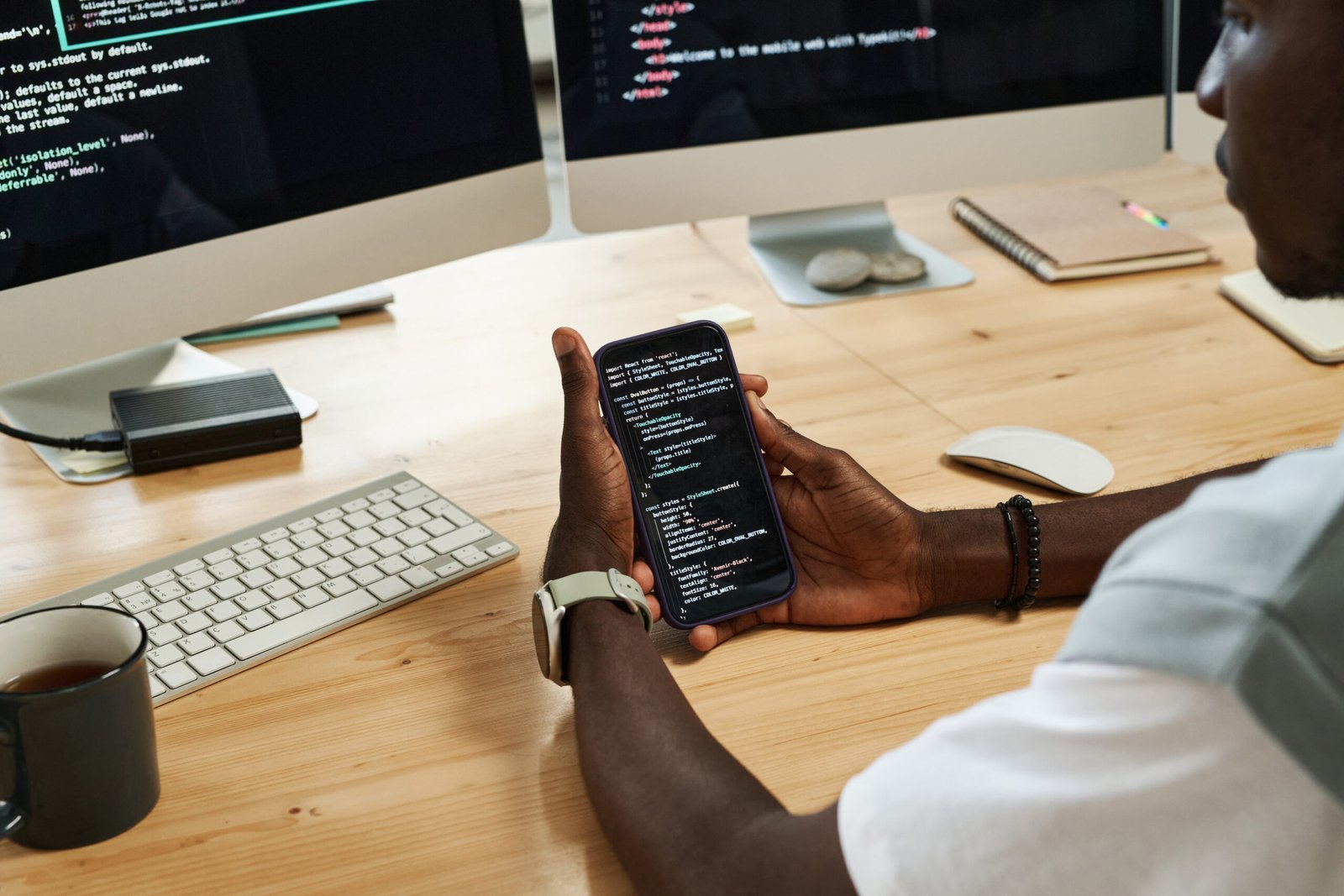 Hands of young black man scrolling through codes in smartphone while sitting by workplace in front of computer screens with data
