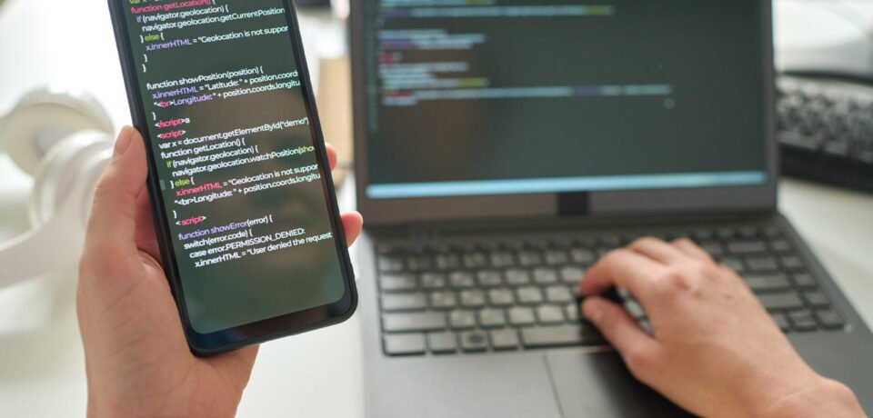 Close-up of programmer holding smartphone with codes in her hands and connecting it with laptop at table
