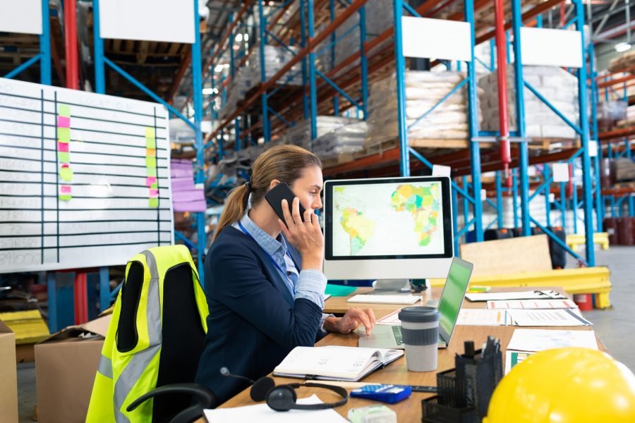 Businesswoman in warehouse talking on phone and working on computer at desk, copy space. Logistics, inventory, distribution, technology, communication, workspace