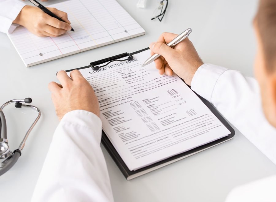Hands of young doctor with pen filling in medical history forms of patients and his assistant or nurse writing down information on blank card
